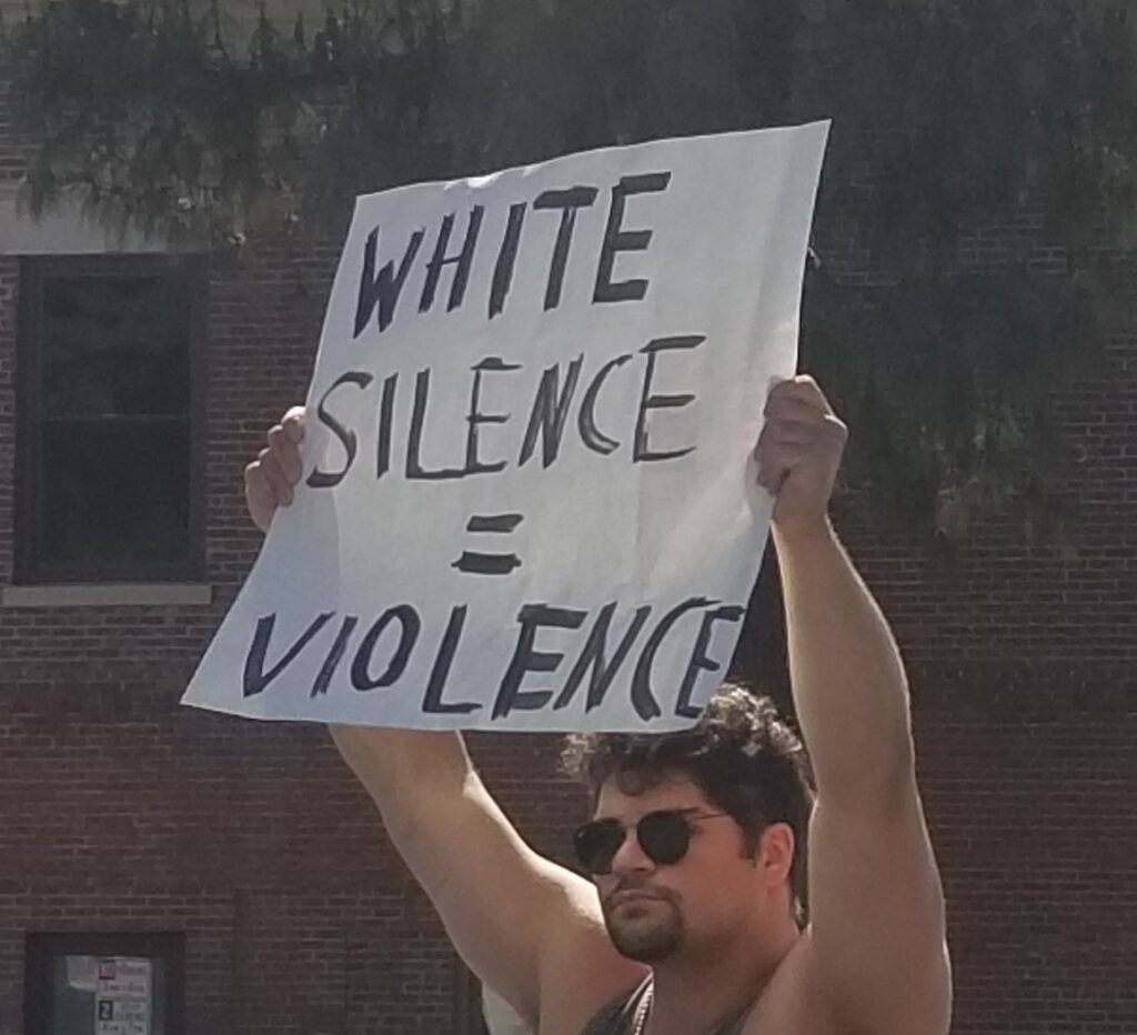 white posterboard sign that reads White silence equals violence. Held by a white man with curly brown hair in sunglasses and a sleeveless shirt.