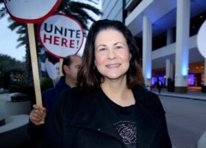 Woman with shoulder length brown hair, smiling. Wearing a dark jacket over a dark t-shirt. Picketers with signs reading "UNITE HERE" in the background