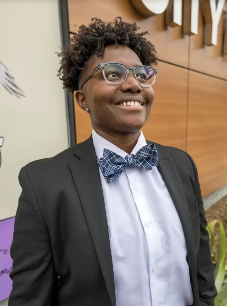 Zekiah Wright, a Black person with natural, short dreads, wearing glasses, smiling and looking up and to the side. Wearing a grey blazer, white button-down shirt and a blue bowtie.