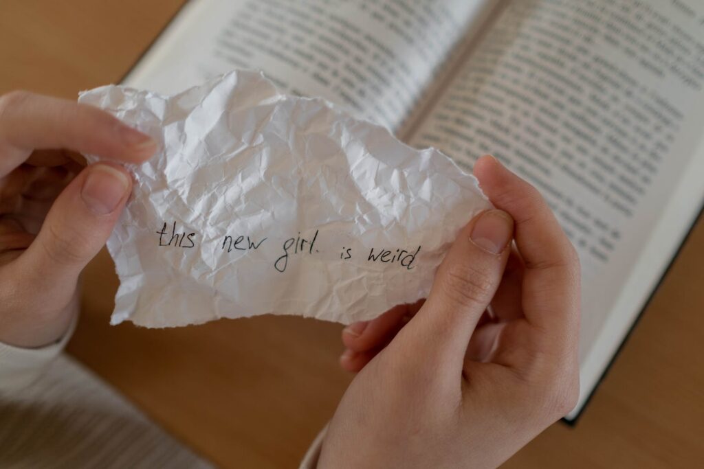 a pair of white hands holding a scrap of paper that reads "This new girl is weird." Background is an open book on a table.