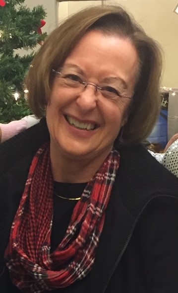 Photo of a white woman smiling. She wears glasses, a black sweater, a red plaid scarf and a gold necklace. Her hair is light brown and a little shorter than shoulder-length.