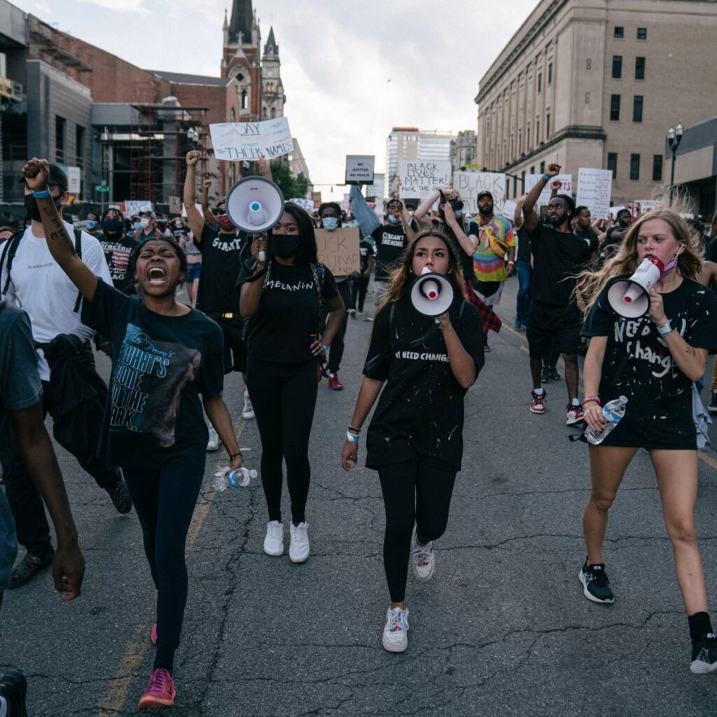 A photo of four teenage girls, wearing black, leading a march down a street in Nashville, Tennessee. Three girls carry bullhorns. One girl's fist is raised in the air and she is shouting. In the background are other marchers carrying Black Lives Matter signs, and city buildings.