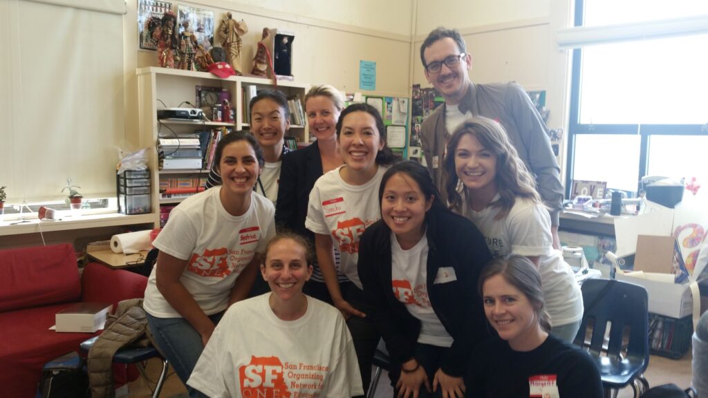 A diverse group of Asian, Latina and white young people smiling. They are wearing white t-shirts with the words SF ONE and a map of San Francisco printed in orange. They are in an elementary school classroom.