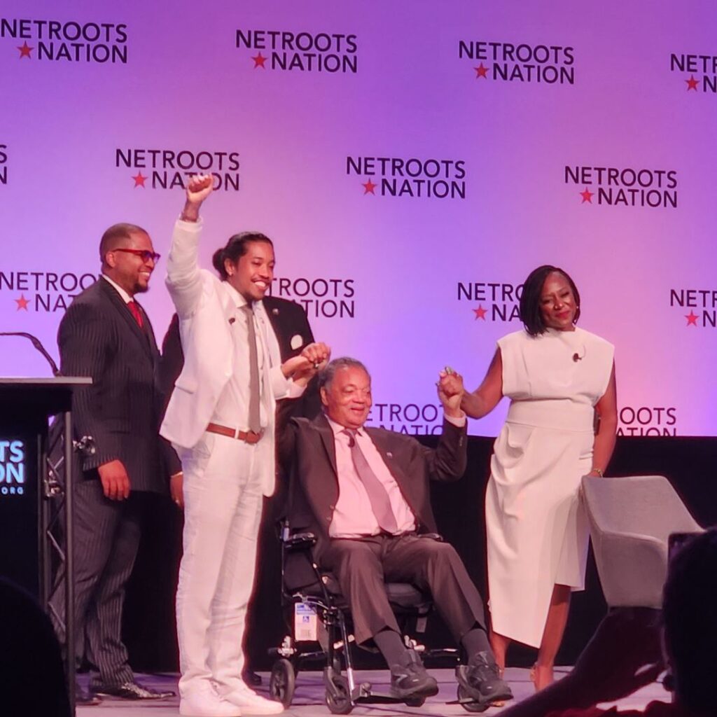 Tennessee state representative Justin Jones, a young, multi-racial man in a white suit, with Alicia Garza, a Black woman in a white suit and Reverend Jesse Jackson, an older Black man in a grey suit, seated in a wheelchair. Jones, Garza and Jackson clasped hands and raised them in victory. Two other Black men in grey suits appear with Jackson. The background is the stage at the Netroots Nation conference.