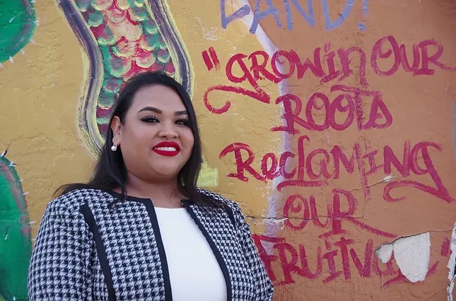 Sasha Ritzie-Hernandez, a young Latinx-Afro-Indigenous woman, smiling, wearing a white shit and black and white checked jacket. She stands in front of an outdoor mural with red lettering that reads Growing our roots reclaiming our Fruitvale. Additional colors in the mural are yellow, brown, green, red and grey.