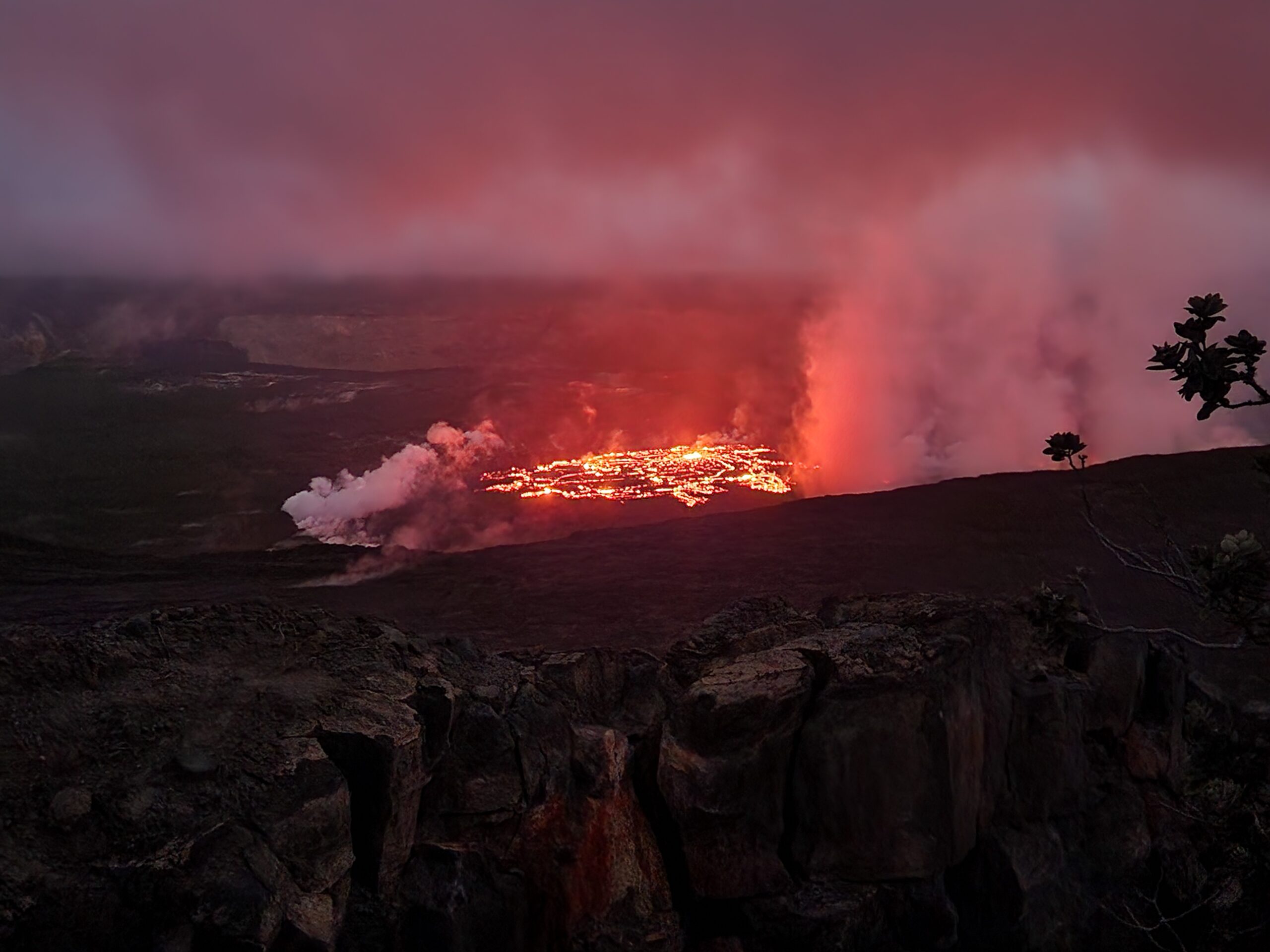 an active volcano seen from the top of an adjacent hill. Red hot lava flows in rivers, surrounded by hills, with steam rising from the edges.