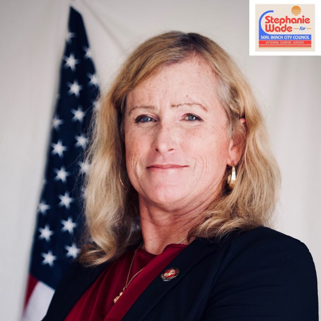 a head shot of a white woman in her mid-fifties, with blond hair, wearing a black jacket and burgundy blouse. There’s a US flag in the background. In the top right corner is a campaign logo for Stephanie Wade for Seal Beach City Council.