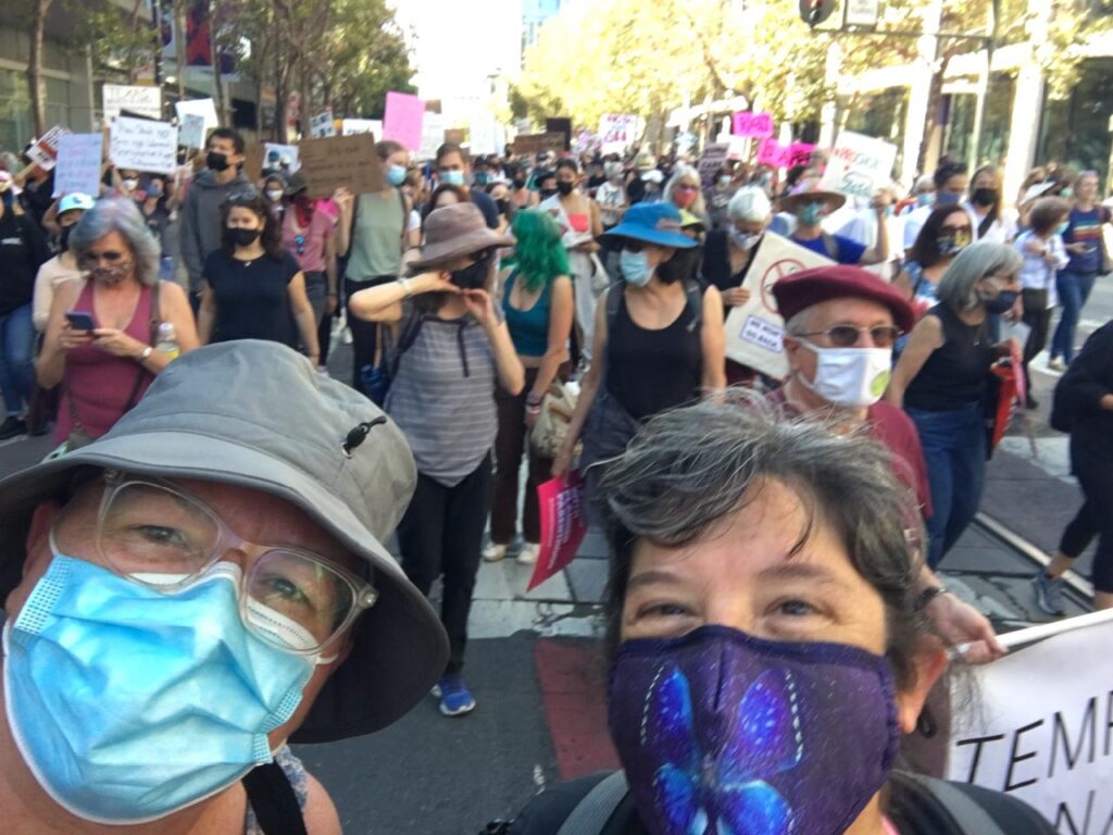 A selfie of two women at an abortion rights march. The woman on the left wears a gray hat, glasses and a blue surgical mask. The woman on the right wears a blue mask with a butterfly.